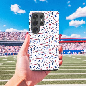 May include: A white phone case featuring a repeating pattern of blue and red footballs, hearts, and stylized buffalo. The case is held in front of a football stadium with a green field and blue sky.