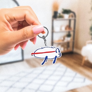 May include: A white keychain with a cartoon illustration of a buffalo, featuring a red stripe and blue accents. The keychain is attached to a silver keyring.