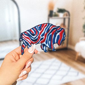 May include: Embroidered patch in the shape of a bison, featuring a red, white, and blue pattern. The patch is held up by a hand with long, manicured nails. The bison patch is a patriotic design.