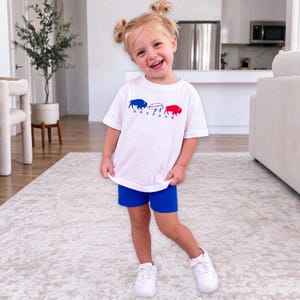 May include: A white short-sleeved t-shirt featuring a graphic of three bison in blue, white, and red, with the word BUFFALO printed in blue letters underneath. The shirt is paired with solid royal blue shorts and white athletic sneakers.