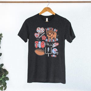 May include: A dark charcoal gray short-sleeve t-shirt featuring a graphic print with the text Let's Go Buffalo and 716. The design includes a cartoon buffalo, a football, a helmet, a heart, a flower, a lightning bolt, and a goal post.