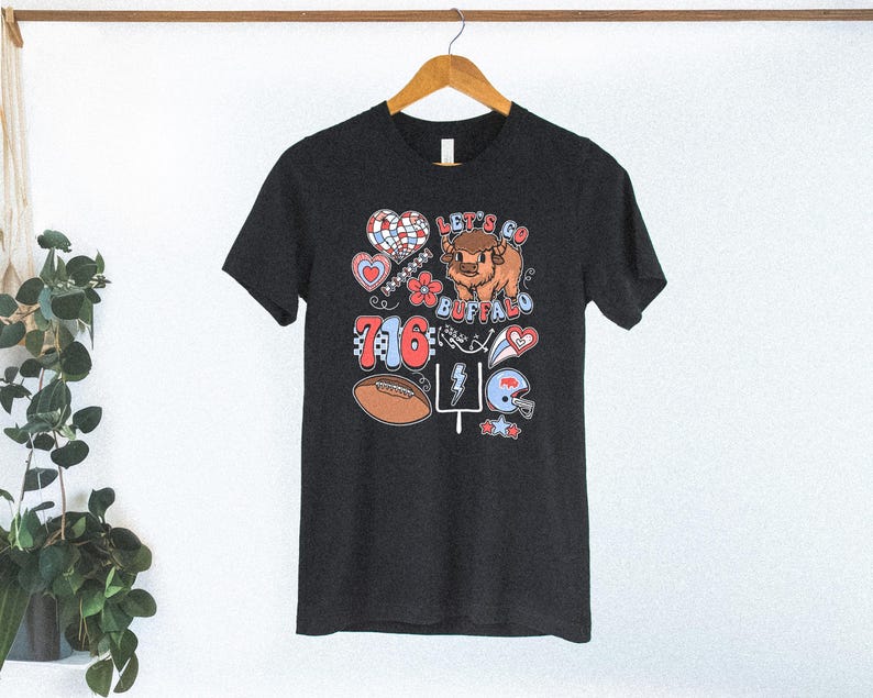 May include: A dark charcoal gray short-sleeve t-shirt featuring a graphic print with the text Let's Go Buffalo and 716. The design includes a cartoon buffalo, a football, a helmet, a heart, a flower, a lightning bolt, and a goal post.
