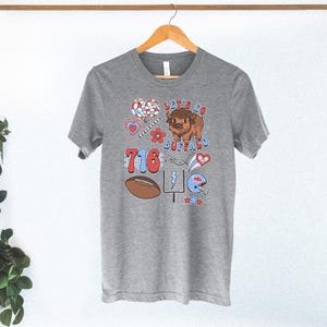 May include: A heather gray short-sleeve t-shirt featuring a graphic print with the text "LET'S GO BUFFALO" and "716". The design includes a brown bison, a football, a goalpost, a blue helmet, hearts, flowers, and stars in red, white, and blue.