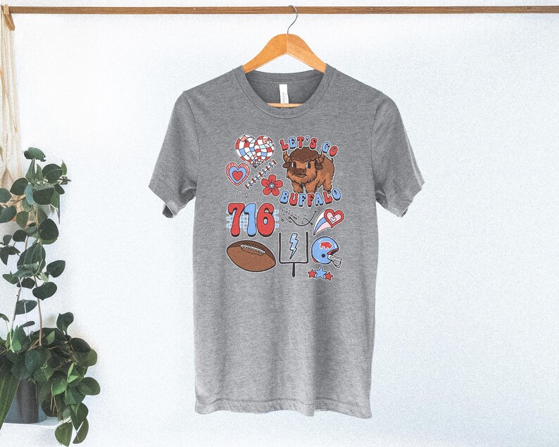 May include: A heather gray short-sleeve t-shirt featuring a graphic print with the text "LET'S GO BUFFALO" and "716". The design includes a brown bison, a football, a goalpost, a blue helmet, hearts, flowers, and stars in red, white, and blue.