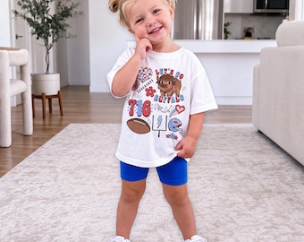 May include: A white short-sleeved t-shirt featuring a graphic print with the text Let's Go Buffalo, a brown bison, a football, a goalpost, a helmet, and the number 716 in red and blue, paired with solid blue athletic shorts and white sneakers.