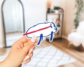 May include: A white sticker featuring a cartoon pig with blue outlines and legs, a red stripe across its body, and two small blue dots. The sticker is held by a hand with manicured nails.