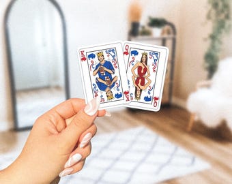 May include: Two playing cards featuring custom illustrations. One card depicts a king in a blue jersey, the other a queen in a red dress. Both cards have a crown and decorative flourishes. The cards are held in a hand with manicured nails.