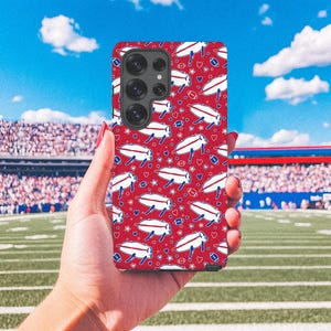 May include: A red phone case with a repeating pattern of white footballs, hearts, and blue and white airplane-like graphics. The case is held in front of a football stadium with a green field and blue sky.