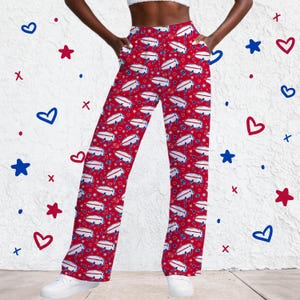 May include: Red lounge pants featuring an all-over repeating pattern of white buffalo silhouettes with blue outlines, small blue footballs, and tiny white stars. These pants have a high-waisted fit, side pockets, and a straight-leg silhouette.