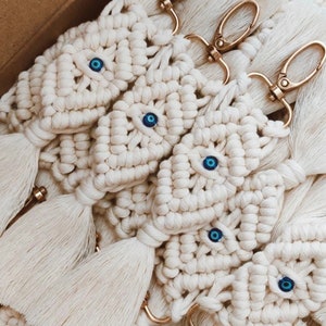 May include: A group of white crocheted keychains with blue evil eye charms and gold key rings. Each keychain has a tassel.