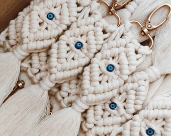 Puede incluir: Un grupo de llaveros de crochet blanco con dijes de ojo azul maligno y anillos de llave dorados. Cada llavero tiene una borla.
