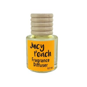 May include: A small, clear glass bottle with a wooden cap and a bright orange label. The label reads "Juicy Peach Fragrance Diffuser" in black script, with "0.2 oz" printed at the bottom. The bottle contains a light yellow liquid.