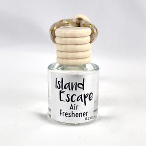 May include: A clear glass bottle with a wooden cap and a gold string hanging from it. The bottle is labeled "Island Escape Air Freshener 0.2 oz."