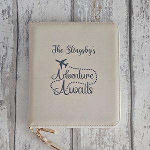 May include: A cream-coloured travel wallet with a gold-tone wrist strap. The front of the wallet features the text "The Singsby's Adventure Awaits" with a small airplane graphic. The wallet is closed and has a zip closure.