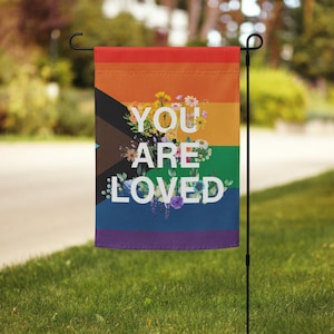 May include: A rainbow flag with the words "You Are Loved" in white lettering surrounded by colorful flowers. The flag is attached to a black metal pole.