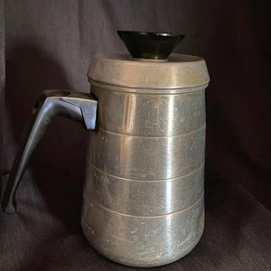 May include: Silver-colored metal coffee percolator with a black lid and a black handle. The percolator has markings for measuring in cups and ounces.