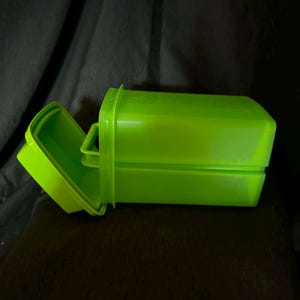 May include: A bright lime green rectangular plastic storage container with a hinged lid. The container features a smooth, solid-colored finish and a secure latch mechanism on the side, designed for holding small items in a compact, stackable form.