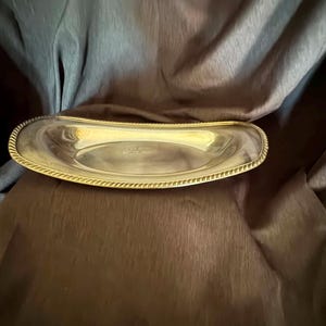May include: An elongated, oval-shaped silver-toned serving tray with a polished, reflective surface. The rim of the tray features a distinct, raised rope-style texture that runs along the entire perimeter, providing a decorative border.