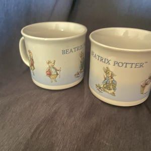 May include: Two white ceramic mugs with blue and white illustrations of Beatrix Potter characters. The mugs have the text "Beatrix Potter" on them.