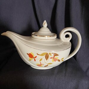 May include: A white teapot with a gold trim and a floral design in shades of orange, yellow, and brown. The teapot has a curved spout and a handle that curls up at the top.