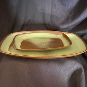 May include: A set of two rectangular ceramic serving trays. The larger tray is a light green color with a brown rim. The smaller tray is a brown color with a light green rim. Both trays have a speckled glaze.