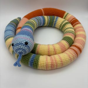 May include: A colorful, crocheted snake toy in a circular shape. The snake features a blue head with black eyes and a blue tongue. The body is striped with orange, pink, yellow, green, and blue. The toy is made of soft yarn.