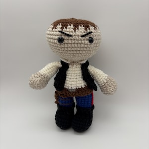 May include: A handmade crochet doll of a character with a light beige head and brown hair. The doll wears a white shirt, black vest, brown belt, blue pants with a red stripe, and black boots. The doll has black eyes and an angry expression.