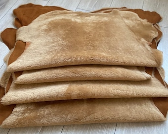 May include: A stack of four brown sheepskin rugs with a soft, fluffy texture. The rugs are folded and arranged on a light wood floor.