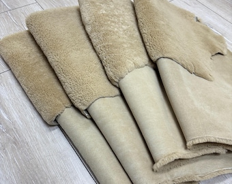 May include: A close-up view of several beige sheepskin pieces, with the soft, textured wool facing up. The underside of the sheepskin is a smooth, light tan color. The pieces are arranged in a layered fashion.