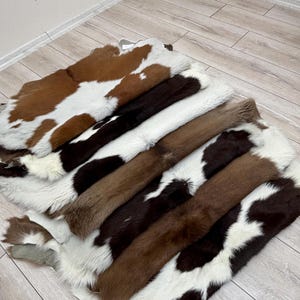 May include: A collection of cowhide rugs in various colors, including brown, white, and black. The rugs are arranged in a layered display, showcasing the natural patterns and textures of the animal hides. The cowhide rugs are a decorative home decor item.