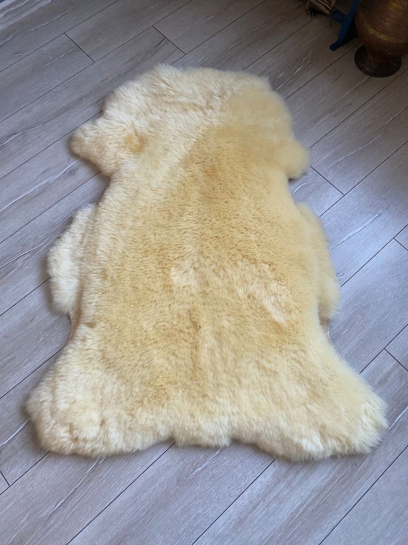 May include: A cream-coloured sheepskin rug with a soft, fluffy texture. The rug is laid out flat on a wooden floor.