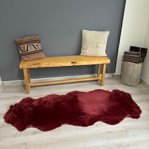 May include: A burgundy faux fur rug with a wavy edge, placed on a light wood floor in front of a wooden bench with two pillows. The bench is against a gray wall.