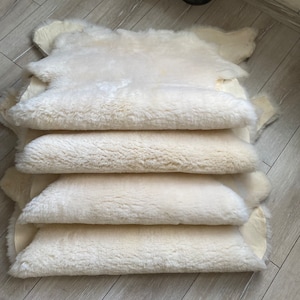 May include: A stack of four off-white sheepskin rugs, with a soft, fluffy texture. The rugs are layered on top of each other, showcasing their natural shape and color. The background is a light-colored wooden floor.