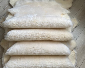 May include: A stack of four off-white sheepskin rugs, with a soft, fluffy texture. The rugs are layered on top of each other, showcasing their natural shape and color. The background is a light-colored wooden floor.