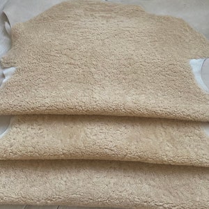 May include: Four beige sheepskin hides stacked on top of each other. The hides are soft and fluffy, with a natural, rustic look.