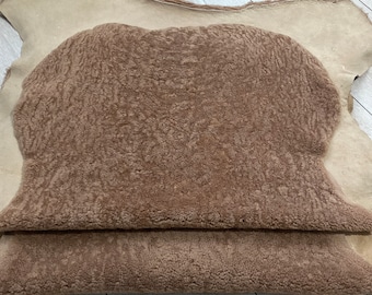 May include: A folded, brown sheepskin rug with a textured surface. The rug is layered over a larger, lighter-colored sheepskin. The image is taken from a high angle, showing the texture and color variations of the natural material.