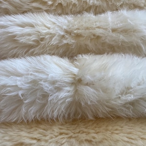 May include: A stack of three fluffy white faux fur rugs. The top two rugs are white and the bottom rug is a light beige colour.