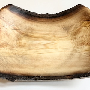 May include: A large, wooden bowl with a natural, light brown finish. The bowl has a slightly irregular shape and a dark brown rim. The wood grain is visible throughout the bowl.