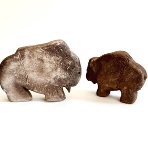 May include: Two carved stone bison sculptures. One is a larger, light gray color with darker gray markings. The other is smaller and a rich brown color. Both are standing and have detailed features.
