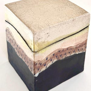 May include: Square decorative box with a textured, off-white lid and sides painted with layers of color. The box features a black base, a brown middle section with blue accents, and a yellow band between the lid and the middle section.