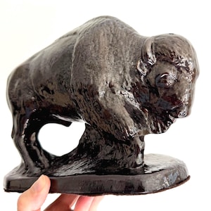 May include: A dark brown, glossy ceramic figurine of a bison standing on an oval base. The sculpture features a textured mane and hump, with a smooth, reflective finish across the body, capturing the animal in a forward-leaning stance.