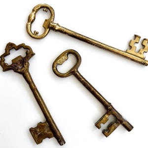 May include: Three antique brass keys of varying sizes and designs are arranged on a white surface. The keys have ornate details, including decorative key heads and intricate teeth. The keys are a warm, aged gold color.