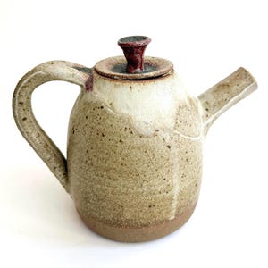 May include: A ceramic teapot with a speckled beige and cream glaze, featuring a curved handle, a straight spout, and a lid topped with a small, dark red knob. The base of the teapot is a solid, unglazed brown clay, contrasting with the textured finish.