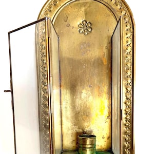 May include: A vintage brass wall-mounted display case with a hinged glass door. The interior features a small brass cup and a decorative flower. The case has an arched top and is adorned with a dotted border.