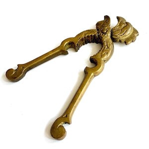 May include: A brass nutcracker featuring a detailed, ornate design with a sculpted head at the hinge and curved, textured handles. The metal has a warm, aged gold tone with visible patina, showcasing intricate carvings along the lever arms.