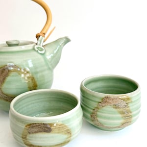 May include: A light green ceramic teapot with a curved rattan handle and a matching lid, accompanied by two small, round ceramic tea cups. Each piece features a subtle horizontal ribbed texture and a decorative brown circular brushstroke pattern.