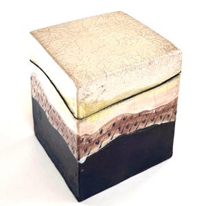 May include: A square ceramic box with a lid featuring a crackled cream-colored top surface. The sides transition from a light cream to a pale pink band with small dark markings, ending in a solid matte black base at the bottom of the container.
