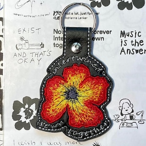 以下が含まれることがあります： 赤と黄色の花の模様の黒い革製キーホルダー。 キーホルダーには「intertwined」の文字と、Adrienne Lenkerの引用句が書かれています。 背景は、絵や手書きのテキストが書かれたノートです。