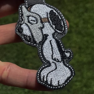 Könnte beinhalten: Ein schwarzer Leder-Schlüsselanhänger mit einem weißen und schwarzen gestickten Cartoon-Hund. Der Hund steht auf seinen Hinterbeinen mit erhobenen Vorderpfoten.