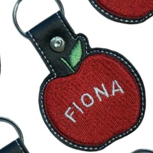 Op de afbeelding: Rode, appelvormige sleutelhanger met de naam "FIONA" in het wit geborduurd. De sleutelhanger heeft een zwarte leren band en een zilveren sleutelring. Het appelontwerp heeft een groen blad.